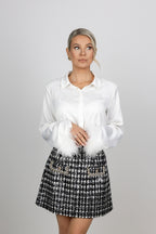 A woman with a white long-sleeve satin blouse with white feather trim at the cuffs, paired with a black and white tweed mini skirt with pearl embellishments