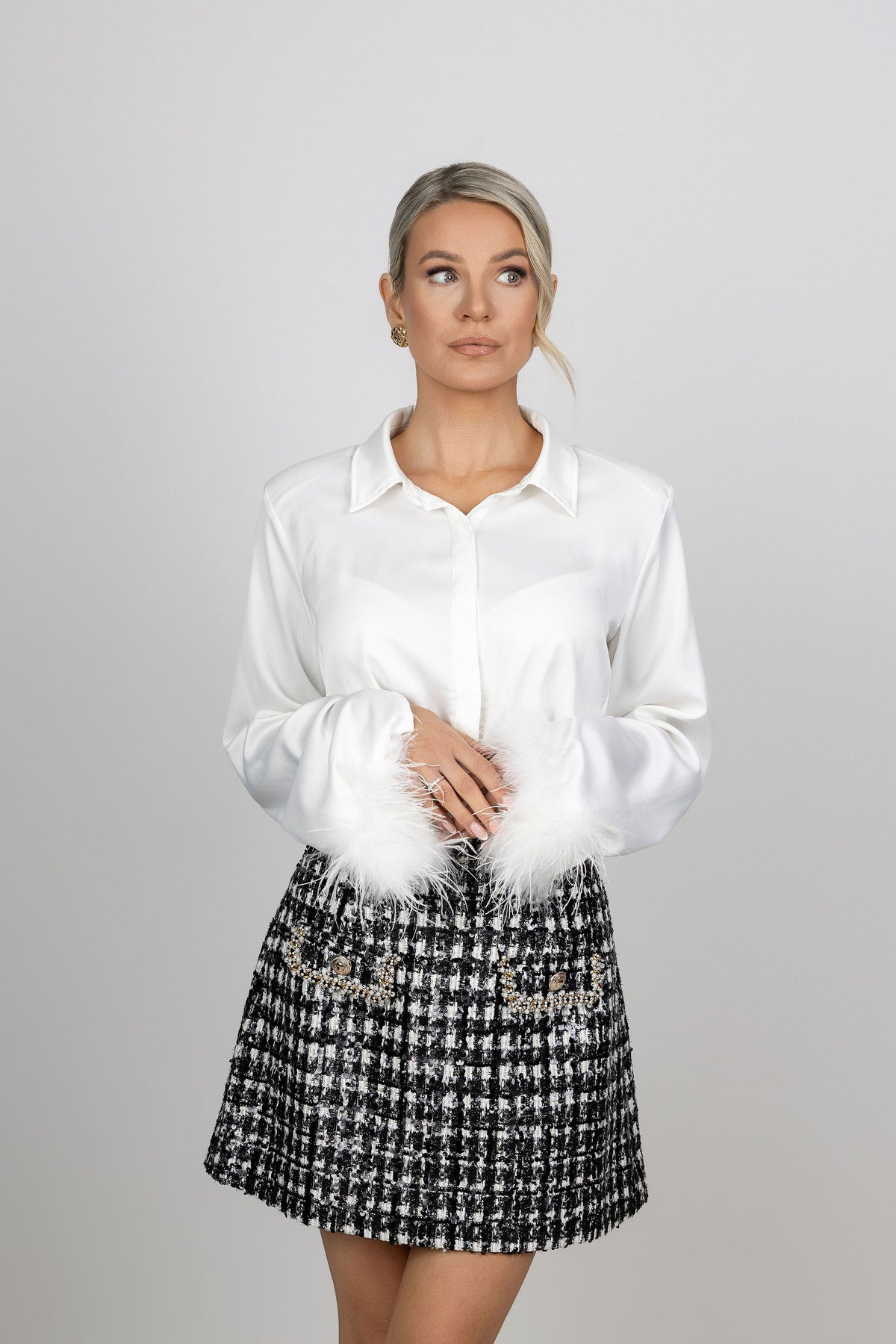 A woman with a white long-sleeve satin blouse with white feather trim at the cuffs, paired with a black and white tweed mini skirt with pearl embellishments