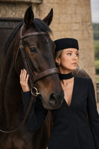 A woman in a black outfit and black pillbox hat stands beside a dark brown horse 